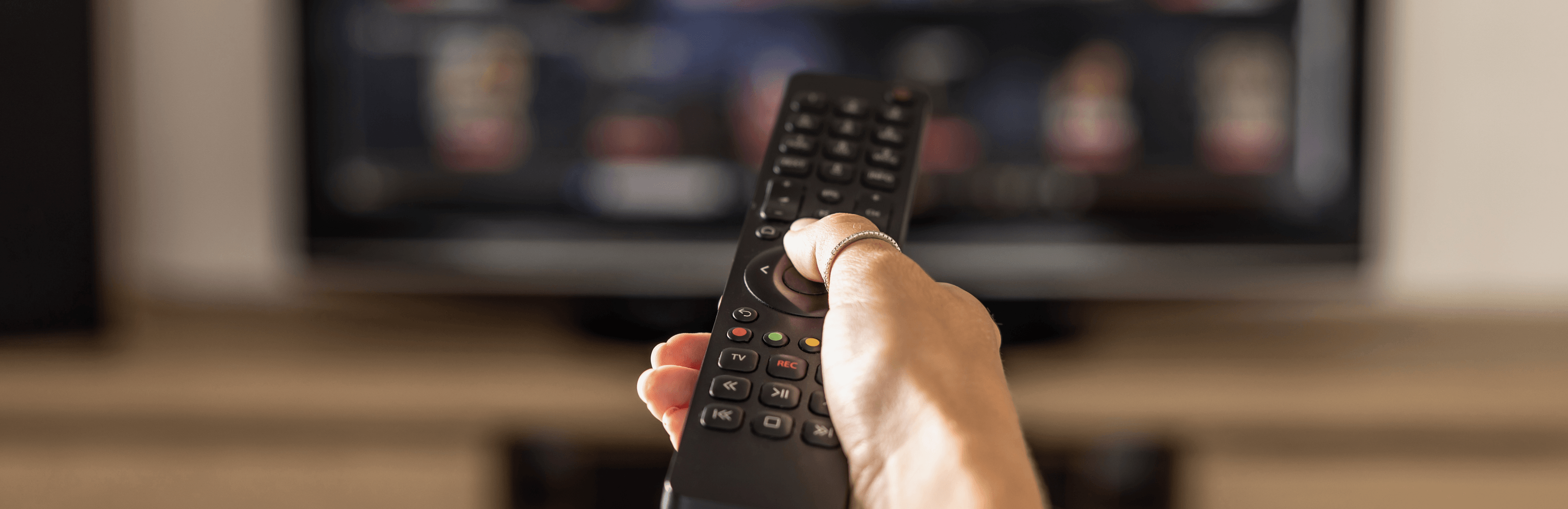 Hand holding a TV remote toward a television, representing the growing complexity of finding and watching live sports across multiple streaming services.